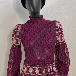 Könnte beinhalten: Ein bordeauxfarbenes Spitzentop mit hohem Rüschenkragen und langen Glockenärmeln. Die Ärmel und die Taille sind mit floralen Stickereien verziert. Auf dem Kopf der Schaufensterpuppe befindet sich ein schwarzer Pelzhut.
