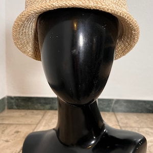 Può includere: Un cappello di paglia intrecciato di colore marrone chiaro con un'ampia tesa. Il cappello è posto su una testa di manichino nera.