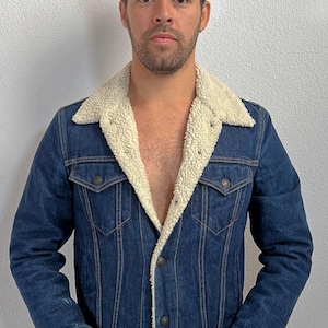 Könnte beinhalten: Dunkelblaue Jeansjacke mit cremefarbenem, flauschigem Shearling-Kragen. Die Jacke hat zwei Brusttaschen und Knopfverschlüsse. Diese Jacke im Vintage-Stil ist ein klassisches Oberbekleidungsstück.