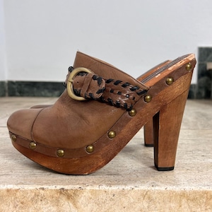 Könnte beinhalten: Braune Lederclogs mit Holzplateau und Absatz. Die Clogs haben einen geflochtenen Riemen mit Schnalle und sind mit messingfarbenen Nieten verziert. Die Absatzhöhe beträgt ca. 10 cm.