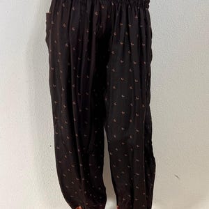 Op de afbeelding: Donkerbruine harembroek met een klein herhalend bloemenpatroon in oranje en geel. De broek heeft een brede, geplooide elastische tailleband en bijpassende elastische boorden bij de enkels, beide afgewerkt met een rood en goudkleurige stoffen rand.