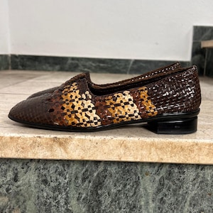 Könnte beinhalten: Ein Paar braune Loafer aus geflochtenem Leder mit kleinem Absatz. Die Schuhe weisen eine Mischung aus dunklen und hellbraunen Tönen in einem Korbgeflechtmuster auf. Die Schuhe werden auf einer Marmoroberfläche präsentiert.