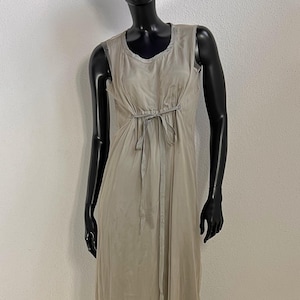 May include: A light beige sleeveless dress with a cinched waist and a tie detail. The dress is made of a sheer fabric and has a flowing, floor-length design. A black hat is on the mannequin's head.