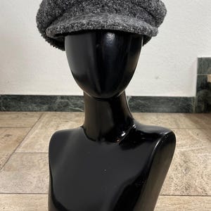 Könnte beinhalten: Graue, strukturierte Ballonmütze, präsentiert auf einem schwarzen Schaufensterpuppenkopf. Die Mütze hat eine runde Krone und einen kleinen, steifen Schirm. Der Hut ist aus weichem, flauschigem Material und hat eine neutrale Farbe.