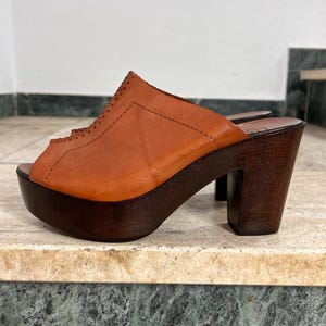 Könnte beinhalten: Braune Leder-Plateausandalen mit offenem Zehenbereich. Die Sandalen haben eine Holzplateausohle und einen klobigen Holzabsatz. Das Obermaterial weist dekorative Nähte auf. Die Schuhe werden auf einer Marmoroberfläche präsentiert.
