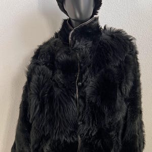 Puede incluir: Una chaqueta negra de manga larga con una textura gruesa y peluda, cuello alto y cierre frontal de botones con detalles de ribetes visibles. La prenda se acompaña de un gorro negro a juego, redondeado y esponjoso, sobre un maniquí.