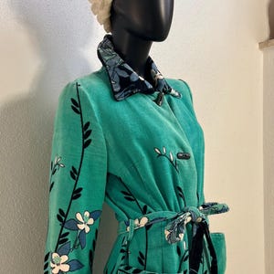 Könnte beinhalten: Ein leuchtend türkisfarbener Cordmantel mit einem wiederkehrenden Blumenmuster aus dunkelblauen und weißen Blüten an langen schwarzen Stielen. Der Mantel verfügt über einen passenden Bindegürtel, zwei Vordertaschen, einen floralen Kragen und Knebelverschlüsse.