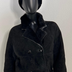 Könnte beinhalten: Schwarze Wildlederjacke mit passendem schwarzem Pelzhut. Die Jacke hat ein doppelreihiges Design mit großen schwarzen Knöpfen, einem Reverskragen und Pelzbesatz an Kragen, Manschetten und Saum. Die Jacke wird auf einer schwarzen Schaufensterpuppe präsentiert.