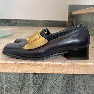 Könnte beinhalten: Ein Paar marineblaue Leder-Loafer mit goldenem Akzent auf dem Spann. Die Schuhe haben eine eckige Zehenpartie und einen kleinen, schwarzen Blockabsatz. Die Loafer werden auf einer Marmoroberfläche präsentiert.