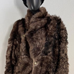 Puede incluir: Una chaqueta de piel marrón de pelo largo y texturizado con cuello alto y botones frontales, exhibida en un maniquí que lleva un gorro negro, redondo y difuso. La chaqueta presenta una apariencia gruesa y peluda con tonos marrones variados.