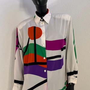 Könnte beinhalten: Weißes Hemd mit farbenfrohem geometrischem Design. Das Hemd hat ein auffälliges Muster aus orangefarbenen, grünen, lila und rosa Formen, die schwarz umrandet sind. Kragen und Manschetten sind weiß. Das Hemd wird auf einer Schaufensterpuppe präsentiert.