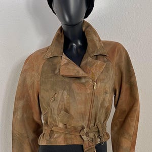 Peut inclure: Veste en daim marron avec fermeture éclair et ceinture. La veste présente un motif moucheté unique et un col classique. Un chapeau en fourrure noire est posé sur la tête du mannequin. La veste est de style vintage.