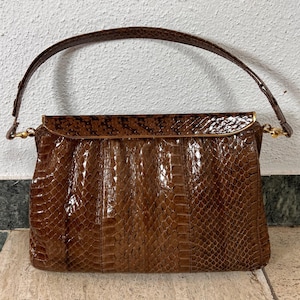 May include: A brown handbag with a glossy, textured snakeskin pattern. The bag features a single arched handle attached with gold-toned metal hardware, a rectangular body with a flap closure, and a structured, polished finish throughout.