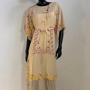 Könnte beinhalten: Ein beigefarbenes, wadenlanges Kleid aus transparentem Lagenstoff mit Bindedetails am Ausschnitt und in der Taille. Das Kleid ist mit geometrischen kastanienbraunen Mustern und gelben kreisförmigen Motiven am Saum und an den Ärmeln verziert, ergänzt durch einen breitkrempigen Hut.