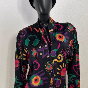 Op de afbeelding: Zwarte blouse met een kleurrijk abstract patroon. Het patroon bevat geometrische en bloemvormen in rood, geel, groen, paars en roze. De blouse met lange mouwen heeft een kraag en wordt gecombineerd met een zwarte bontmuts.