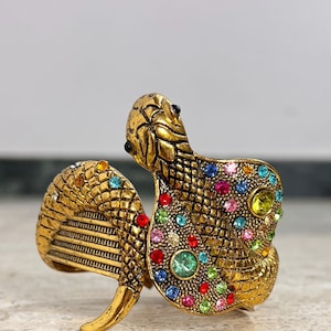 Può includere: Bracciale a polsino a forma di serpente color oro con accenti di pietre preziose colorate. Il bracciale presenta un design dettagliato a forma di serpente con una finitura strutturata e una varietà di pietre preziose rotonde colorate.