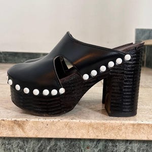 Könnte beinhalten: Schwarze Plateau-Clogs mit Peep-Toe-Design. Die Clogs haben einen dicken, strukturierten Absatz und eine Plateau-Basis, verziert mit weißen runden Nieten. Das Obermaterial besteht aus glattem, schwarzem Material. Absatzhöhe ca. 10 cm.