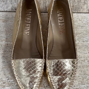 Könnte beinhalten: Ein Paar goldfarbene Ballerinas mit einem strukturierten, schlangenartigen Muster auf der Spitze und den Seiten. Das Wort "MADELEINE" ist goldfarben in jeden Schuh gedruckt. Die Schuhe haben eine spitze Zehenpartie und eine genähte Zierleiste.