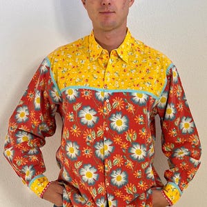Puede incluir: Una camisa de manga larga con botones que presenta una base roja vibrante con un gran estampado floral blanco y azul. La parte superior del pecho y el cuello son amarillos con un pequeño estampado floral blanco, separados por un ribete azul claro.