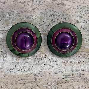 May include: A pair of circular, vintage earrings. Each earring has a deep purple centre, encircled by a pink ring and a green outer ring. A thin gold-coloured metal band separates the colours. The earrings are set against a speckled background.