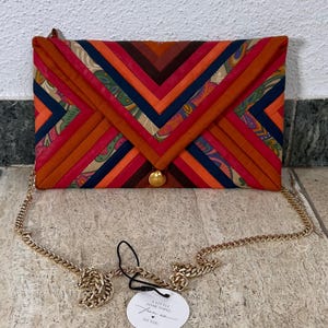Op de afbeelding: Een kleurrijke clutch met een gouden kettingriem. De tas heeft een geometrisch ontwerp met oranje, rode en blauwe stoffen panelen. Een kleine goudkleurige sluiting zekert de flap. Een label leest "A little something from us to you."