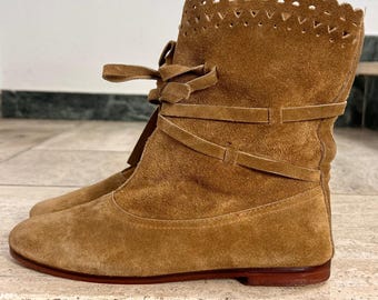 May include: A pair of brown suede ankle boots. The boots feature a scalloped top edge and decorative ties. The low-heeled boots have a rounded toe and a reddish-brown sole. The boots are made of a soft, textured material.