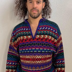 Könnte beinhalten: Ein farbenfroher Pullover mit geometrischem Muster in Rot, Blau, Grün und Braun. Der Pullover hat einen V-Ausschnitt und lange Ärmel. Die Bündchen und der Halsausschnitt sind blau.
