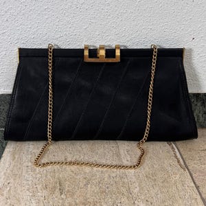 Könnte beinhalten: Schwarze Clutch mit goldfarbener Kette und Akzenten. Die rechteckige Tasche hat ein gestepptes Design auf der Vorderseite und einen goldfarbenen Verschluss. Der Kettenriemen ist an den Seiten der Tasche befestigt.