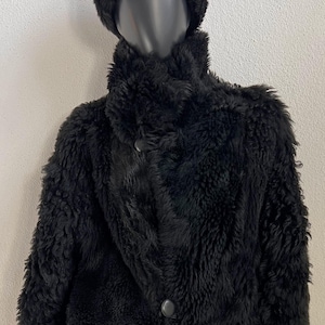 Könnte beinhalten: Eine schwarze Kunstfelljacke mit hohem Kragen und zwei sichtbaren Knöpfen, kombiniert mit einer passenden schwarzen Kunstfellmütze. Die Jacke hat lange Ärmel mit glatten schwarzen Bündchen und eine strukturierte, zottelige Oberfläche.