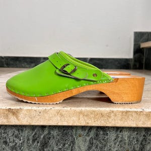 Könnte beinhalten: Hellgrüner Holzschuh mit Holzsohle und Riemen mit Schnalle. Der Schuh hat eine runde Spitze und einen kleinen Absatz. Das Obermaterial ist leuchtend grün, die Holzsohle hellbraun.