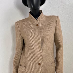 May include: A tan and white diagonal striped blazer featuring a high collar, long sleeves, and two brown buttons. The jacket has a structured shoulder design and two front pockets with flaps, presented on a black mannequin with a black faux fur hat.