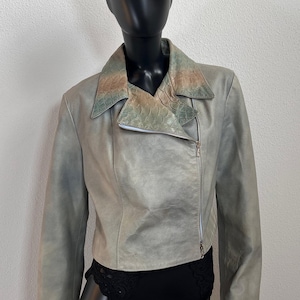 Könnte beinhalten: Hellgraue Lederjacke mit asymmetrischem Reißverschluss, kombiniert mit einer gemusterten Ballonmütze. Die Jacke hat einen strukturierten Kragen mit einem reptilienartigen Muster in Grün- und Brauntönen.