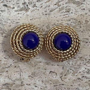 Könnte beinhalten: Ein Paar Clip-Ohrringe mit einem goldfarbenen, seilartigen Design, das einen tiefblauen, kreisförmigen Mittelpunkt umgibt. Die Ohrringe sind vor einem neutralen, strukturierten Hintergrund platziert.