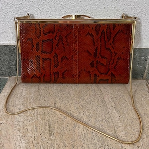 Könnte beinhalten: Eine Vintage-Clutch-Tasche mit Schlangenmuster in Braun- und Schwarztönen. Die rechteckige Tasche hat einen goldfarbenen Rahmen und einen zarten Kettenriemen. Die Tasche ist ein klassisches Accessoire, das sich für formelle Anlässe oder den täglichen Gebrauch eignet.