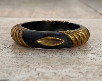 May include: A black wooden bangle with gold metal accents. The bangle has a decorative, diamond-shaped design in the center.
