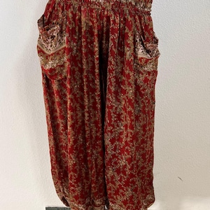 Könnte beinhalten: Weit geschnittene Hose in einem tiefen Rotton mit einem komplexen floralen Muster in Gold und Braun. Das Kleidungsstück verfügt über einen breiten, gesmokten Gummibund, zwei Seitentaschen und geraffte Beinabschlüsse für eine voluminöse Form.