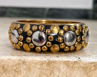 May include: A wide, black bangle bracelet with gold and silver embellishments. The bracelet features a series of small gold and silver circular studs, interspersed with larger silver and gold circular accents. The interior of the bracelet is gold.