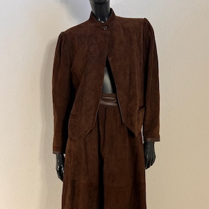 Könnte beinhalten: Ein zweiteiliges Set aus dunkelbraunem Wildleder, bestehend aus einer langärmeligen Jacke mit Stehkragen und einem Knopfverschluss sowie einem passenden wadenlangen Rock mit glattem Bund. Das Set wird auf einer schwarzen Schaufensterpuppe präsentiert.