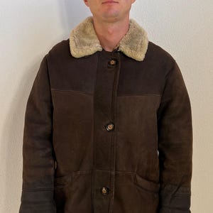 Puede incluir: Un abrigo de ante marrón oscuro con cuello de borreguillo color crema, cierre frontal con cuatro botones oscuros visibles, mangas largas con puños reforzados y dobladillo recto. El abrigo presenta una textura suave y un corte estructurado.