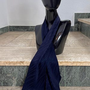 Könnte beinhalten: Ein marineblauer Schal mit geripptem, strukturiertem Design, drapiert über einer schwarzen Mannequin-Büste. Der Stoff des Schals hat einen dezenten Glanz, und die Enden sind spitz. Die Büste befindet sich auf einer Steintreppe.