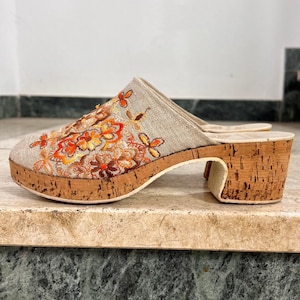 Op de afbeelding: Een instap-muiltje met een beige linnen bovenwerk en gedetailleerd bloemenborduursel in oranje, geel en bruin. De schoen is voorzien van een dikke, gestructureerde kurken plateauzool en een blokhak met een natuurlijke, lichtbruine afwerking.