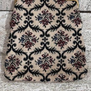 Könnte beinhalten: Vintage Clutch aus beigefarbenem Gobelin mit goldfarbenem Verschluss. Der Stoff hat ein sich wiederholendes Muster aus schwarzen, burgunderroten und blauen Blumenmotiven. Die Tasche hat eine strukturierte Form und ein klassisches, zeitloses Design.