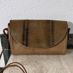 May include: A rectangular shoulder bag featuring a smooth tan leather body and a curved flap with a textured, dark brown snakeskin-patterned overlay. The bag includes thin, matching tan leather shoulder straps attached to the sides.
