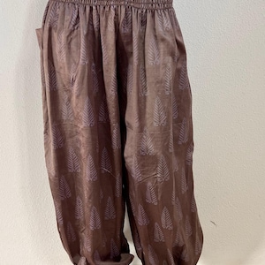 May include: Brown harem pants featuring a repeating light brown fern leaf pattern. The garment has a wide, gathered elasticized waistband and loose, billowy legs that taper at the ankles with elastic cuffs. The fabric has a subtle sheen throughout.