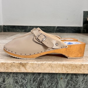 Könnte beinhalten: Beige Clogs mit Holzsohlen. Die Clogs haben einen Lederriemen mit silberner Schnalle und ein beiges Obermaterial aus Leder. Die Holzsohle ist hellbraun. Die Clogs stehen auf einer Marmoroberfläche.