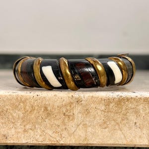 Pode incluir: Uma pulseira artesanal com segmentos alternados de madeira escura, osso branco e metal cor de latão. A pulseira tem uma forma curva e foi concebida para ser usada no pulso. Os materiais criam um contraste marcante.