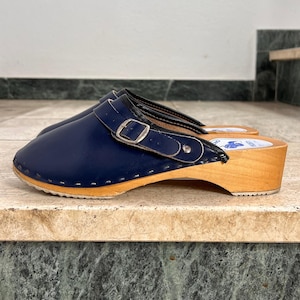 Könnte beinhalten: Ein Paar marineblaue Clogs mit Holzsohlen. Die Clogs haben ein Obermaterial aus Leder mit Schnallenverschluss und eine runde Spitze. Die Holzsohle ist hellbraun. Die Clogs sind ein klassischer Schuhstil.