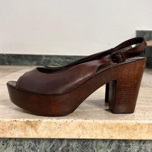Könnte beinhalten: Braune Leder-Plateausandale mit offenem Zehenbereich. Die Sandale hat eine Holzplattform und einen Blockabsatz. Die Absatzhöhe beträgt ca. 10 cm. Die Sandale hat einen Slingback-Riemen mit Schnallenverschluss.