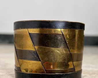 May include: A cylindrical cuff bracelet featuring a geometric pattern of alternating brass and dark metal segments. The design includes solid dark bands at the top and bottom edges, with a series of angled, interlocking metal pieces in the center.