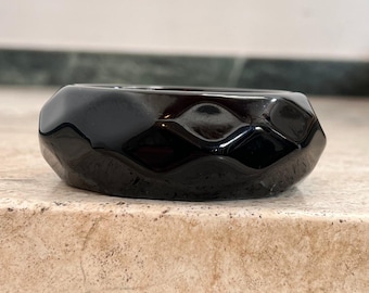 May include: A black, faceted, glass bangle bracelet. The bracelet is shiny and has a geometric design.